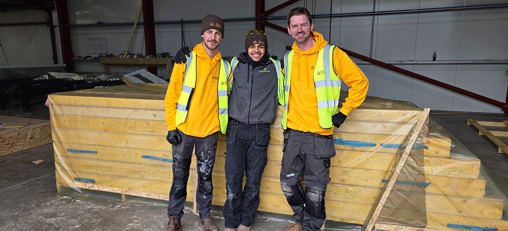 Three Fleming Homes team members standing beside wrapped timber frame panels in the workshop